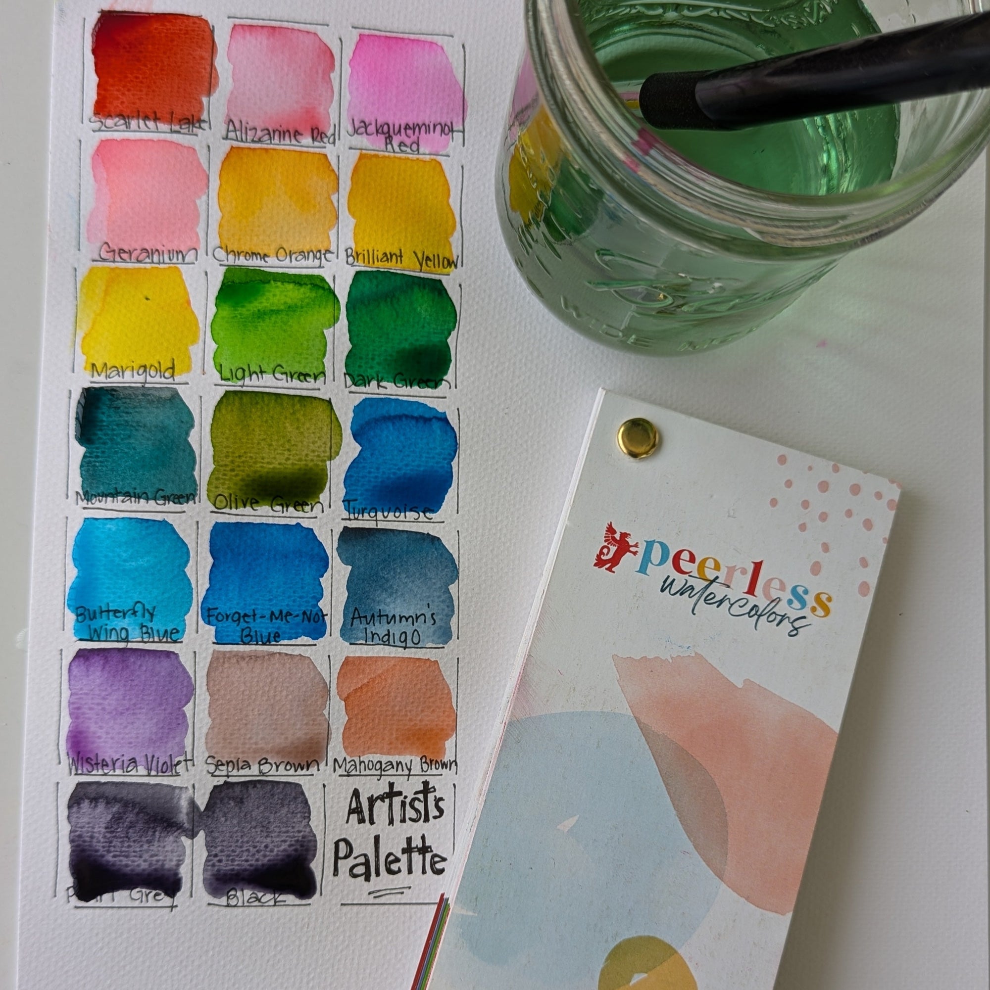 Watercolor paint palette with a jar of water and a color swatch book on a white surface.